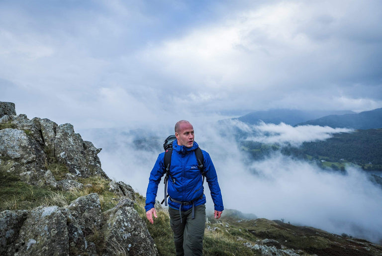 A man walking up a mountain, above clouds in the background and wearing a blue waterproof jacket and a backpack - action