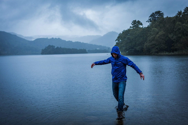 A man walking over stepping stones by a lake on a rainy day and wearing a blue waterproof jacket with the hood up - action