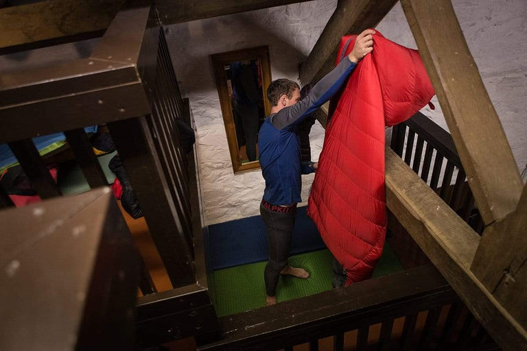 A man wearing merino wool base layers and hanging his red sleeping bag over a wooden beam in a barn - action