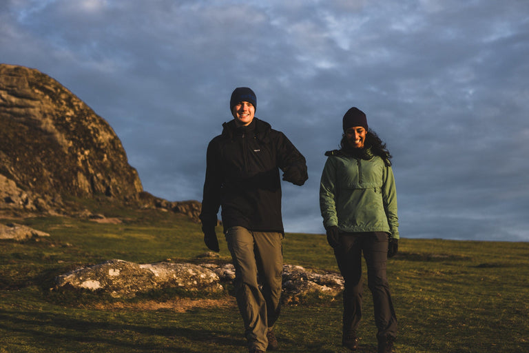 A man and a woman walking at dawn on the Devon coast, wearing black and green Jura Mountain Smocks- action - closed