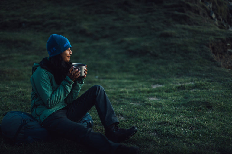 A woman drinking a hot drinking and watching the sunset, sat on her rucksack, wearing a green Jura Mountain Smock - action