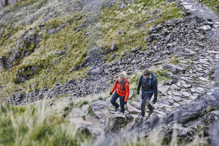 A man and women wearing waterproof clothing with backpacks on walking up a rocky path - action|apy